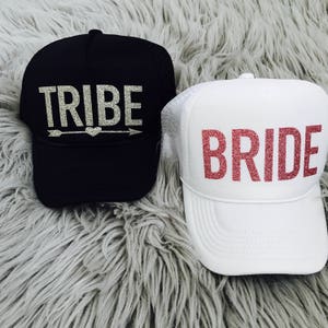May include: Two trucker hats, one black with silver glitter text that says "TRIBE" with an arrow and heart, and one white with pink glitter text that says "BRIDE".