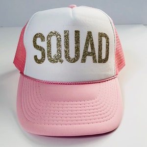 May include: A pink and white trucker hat with a gold glitter "SQUAD" design.