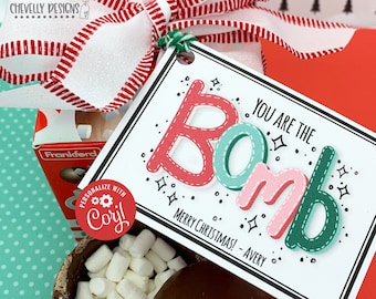Puede incluir: Una caja de regalo con una cinta a rayas rojas y blancas y una etiqueta que dice "You are the Bomb". La etiqueta tiene letras rojas, verdes y rosas. Se ve una taza de malvaviscos y una bomba de chocolate caliente.