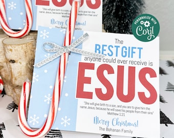 May include: A red and white candy cane with a silver ribbon tied around it. The candy cane is placed on a blue and white card with the text "The Best Gift anyone could ever receive is JESUS" in red and white letters. The card also includes a Bible verse from Matthew 1:21 and the text "Merry Christmas! The Bohanan Family".