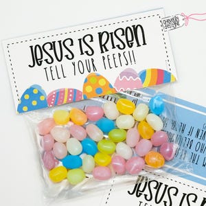 Puede incluir: Una tarjeta blanca con el texto "Jesús ha resucitado, ¡díselo a tus amigos!" impreso en negro. La tarjeta está decorada con huevos de Pascua coloridos y está unida a una bolsa de plástico transparente llena de gominolas de colores pastel.