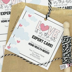 May include: A white tag with pink and black text that reads "Love is in the air and so is our expert care!" with a pink heart and blue clouds. The tag is tied with black and white twine and has a small brown paper bag behind it. The tag also includes the text "Give us a call for all of you home health needs!" and contact information.
