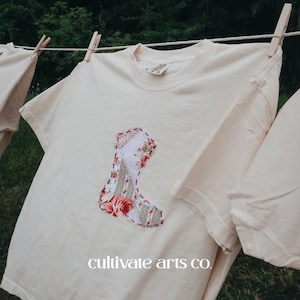 May include: Cream-colored t-shirt with a floral patchwork boot design. The boot features red roses and green leaves on a white and gray background. The text "cultivate arts co." is printed at the bottom of the shirt.