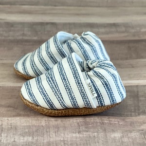 Puede incluir: Un par de zapatos de bebé a rayas blancas y azules con suela marrón. Los zapatos tienen un detalle anudado y una etiqueta que dice "TWINS".