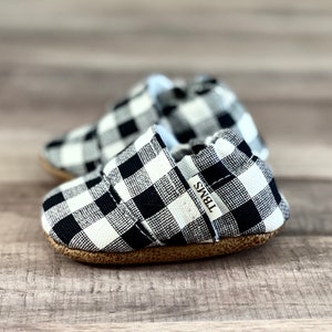 以下が含まれることがあります： 黒と白のチェック柄のベビーシューズのペア。茶色のソール。シューズには「TBMS」と書かれたラベルが付いています。