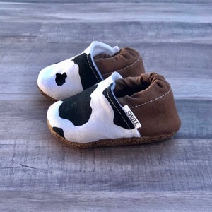 May include: A pair of brown and white cow print baby shoes with a brown fabric cuff and the word "TIBBS" printed on the side.