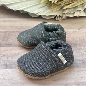May include: A pair of black canvas baby shoes with a brown sole. The shoes have a white label on the side that says "Made with Love".