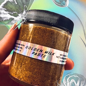 Op de afbeelding: Een glazen pot gevuld met een bruine gouden melkpasta. De pot heeft een zwarte deksel en een holografisch etiket met de tekst "Golden Milk Paste". Het etiket bevat ook gebruiksaanwijzingen: "voeg een theelepel toe aan NIET kokend water, enzymen &"