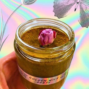 Op de afbeelding: Een glazen pot gevuld met een goudbruine pasta met het label "Golden Milk Paste". Een gedroogde roze rozenknop ligt bovenop de pasta.