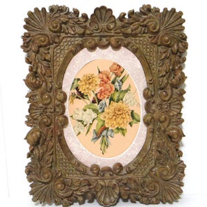 May include: A rectangular picture frame with a dark brown, ornate, carved border featuring scrollwork and shell motifs. The frame surrounds an oval-shaped print of a colorful floral bouquet with yellow, orange, and white flowers on a light background.