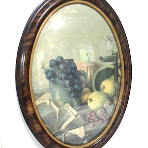 May include: Oval framed still life art print featuring a collection of fruits, including grapes, apples, and a pumpkin. The artwork is set against a neutral background, with a dark wooden frame and a gold inner border. The print also includes a book and a metal pitcher.