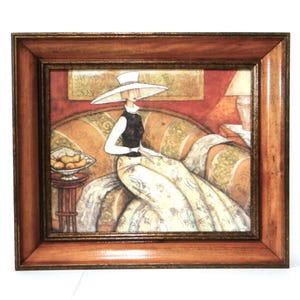 Könnte beinhalten: Gerahmtes Kunstwerk, das eine Frau mit weißem Hut und gemustertem Kleid zeigt, die auf einem Sofa sitzt. Das Kunstwerk hat eine warme Farbpalette und einen braunen Holzrahmen. Eine Schale mit Obst steht auf einem kleinen Tisch.