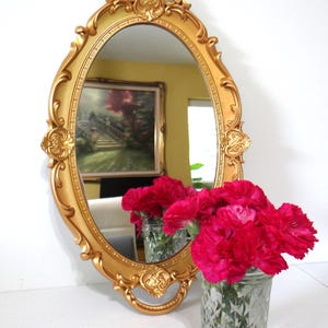May include: An ornate, gold-colored oval mirror with decorative detailing. The mirror reflects a painting and a bouquet of bright pink carnations in a clear glass jar. The mirror is a decorative home decor item.