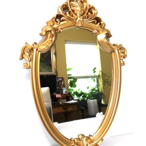 May include: Ornate gold-colored mirror with an oval shape. The mirror has a decorative frame with intricate scrollwork and floral accents. The mirror reflects a room with a window and plants.