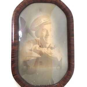 May include: An oval-shaped, vintage photograph in a dark brown wooden frame. The sepia-toned image features a smiling person in a military cap and uniform. The frame has a glossy finish and rounded corners, adding a classic touch.
