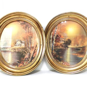 May include: Two oval framed landscape prints. Each print depicts a house near water with trees. The gold-coloured frames have a beaded edge. The artwork uses a warm colour palette, with autumnal tones.