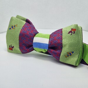 Pode incluir: Uma gravata borboleta colorida com um design único. A gravata borboleta apresenta uma seção central com um tecido bordô estampado, ladeada por seções verde claro com bordados de peixes. A faixa central tem listras verdes, brancas e azuis.