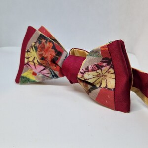 May include: A handmade bow tie with a colorful floral pattern. The bow tie features a burgundy center and edges, with a mix of red, yellow, and beige floral designs. The bow tie is set against a white background.