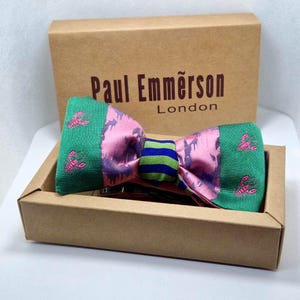 Pode incluir: Uma gravata borboleta colorida com um design rosa e verde, com motivos de lagosta bordados. A gravata borboleta é apresentada numa caixa de cartão castanha com o texto "Paul Emmerson London". A gravata borboleta tem um centro listrado.