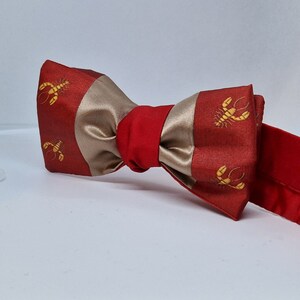 Pode incluir: Uma gravata borboleta vermelha e bege com um padrão de lagosta. A gravata borboleta tem um centro vermelho e extremidades vermelhas, com uma seção central bege. As seções vermelhas têm um padrão repetido de lagostas douradas.