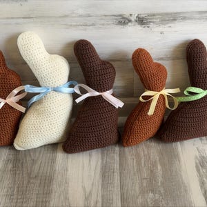 May include: Five handmade crochet bunny rabbits in shades of brown and cream, each with a ribbon tied around its neck. The ribbons are in various colors, including pink, blue, and yellow. The bunnies are arranged on a light wood-grain surface.