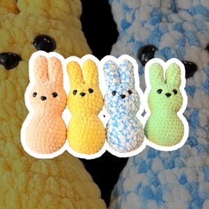 May include: Four handmade crochet bunny figures in pastel colors: peach, yellow, blue, and green. Each bunny has black bead eyes and a rounded body with upright ears. The background features larger, out-of-focus yellow and blue crochet textures.