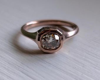 Op de afbeelding: Een roségouden ring met een vierkant geslepen, lichtgekleurde edelsteen. De ring heeft een eenvoudige band en een bezel setting. De ring wordt gepresenteerd op een wit oppervlak.
