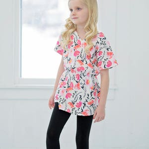 May include: A young person wearing a white and pink floral print top with a black elastic waistband, black leggings, and white ballet flats. The top has a V-neck and short sleeves. The person is standing in front of a white wall with a window in the background. The image includes the text "simplelife PATTERN COMPANY".