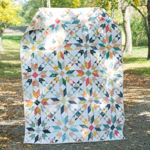 May include: A handmade quilt with a starburst pattern in a variety of colors, including white, blue, orange, and green. The quilt is displayed outdoors, with a backdrop of trees and a gravel path.