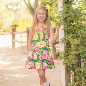 May include: A young girl wearing a green, yellow and pink floral dress with a white collar and yellow buttons. She is standing next to a white wooden fence with green vines growing on it. She is wearing white socks and brown boots with pink laces.