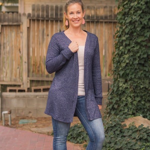 Può includere: Una donna indossa un cardigan blu sopra una canottiera bianca e jeans blu. Indossa scarpe con tacco marroni e orecchini rosa.