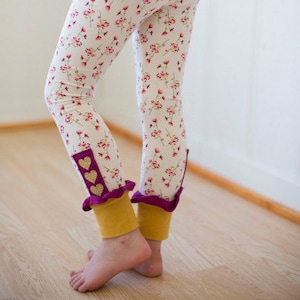 Puede incluir: Leggings blancos con estampado floral y detalles de corazones dorados en los puños. Los leggings tienen un ribete de volantes morado.