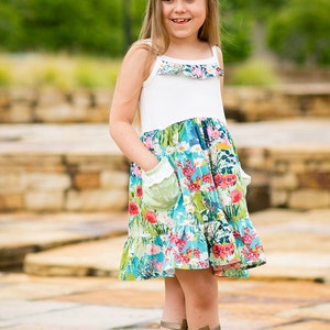 May include: A young girl wearing a white and floral print dress with a green ruffle trim and a flower crown. She is wearing brown gladiator sandals and standing on a stone path.