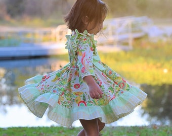 Könnte beinhalten: Ein junges Mädchen in einem grün-weiß gemusterten Kleid mit Rüschen am Saum, das auf einer grünen Wiese steht. Sie trägt goldene Schuhe und hat ihre Haare nach hinten gebunden.