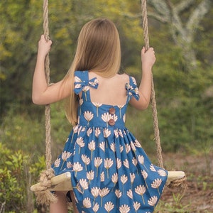 May include: A young girl wearing a blue and white floral dress with a bow on each shoulder strap. She is sitting on a swing in a wooded area.