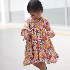 Könnte beinhalten: Ein junges Mädchen trägt ein weißes Kleid mit einem bunten Herzmuster. Das Kleid hat einen Rüschen-Saum und Glockenärmel. Sie trägt schwarze Schuhe mit rosa Schleifen.