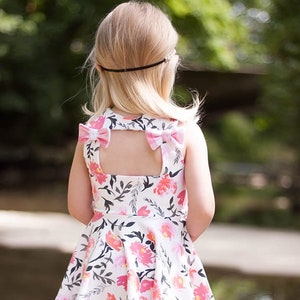 May include: A white sleeveless dress with a floral print and pink bows on the back. The dress has a keyhole opening in the back and a flared skirt. The model is wearing white shorts underneath the dress.