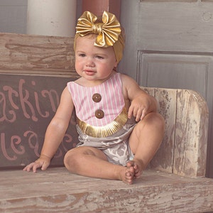 Könnte beinhalten: Ein kleines Mädchen trägt einen rosa-weiß gestreiften Strampler mit goldenen Verzierungen und einem goldenen Schleifen-Haarband. Der Strampler hat zwei große braune Knöpfe vorne. Das Baby sitzt auf einer Holzbank.