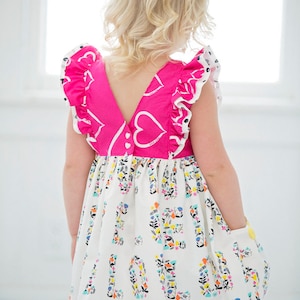Puede incluir: Un vestido rosa y blanco con un patrón de corazón en el corpiño y un estampado floral con la palabra "LOVE" repetida en la falda. El vestido tiene una espalda en forma de V y volantes en las mangas.