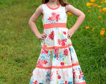 May include: A young girl wearing a white floral dress with a coral sash and a flower crown. She is standing in a field of yellow flowers.