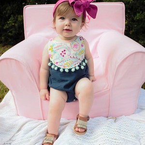 Könnte beinhalten: Ein kleines Mädchen in einem blau-weiß gemusterten Strampler mit einem rosa Schleifen-Haarband sitzt auf einem rosa Sessel. Das Baby trägt braune Sandalen und schaut in die Kamera.