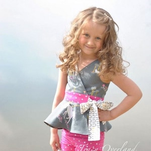 May include: A young girl wearing a grey and white patterned wrap top with a pink and white polka dot sash. The top features a unicorn design. She is also wearing pink and white polka dot shorts. The girl is standing in front of a body of water.