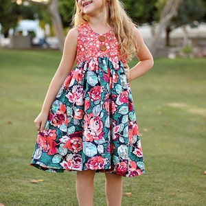 May include: A young girl wearing a sleeveless floral dress with a coral bodice and a coral flower headband. She is standing on a grassy lawn.