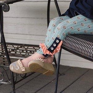 Puede incluir: Una persona joven lleva pantalones de punto azules y verdes con un volante de color melocotón y un detalle de botones negros. Los pantalones de punto se combinan con bailarinas beige con un detalle de lazo plateado. La persona está sentada en una silla de metal negro.