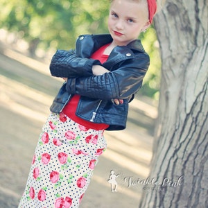 May include: A young girl wearing a black leather jacket, a red shirt, and a white skirt with pink roses and blue polka dots. She has her hair styled in a vintage updo with a red headband. The image is taken outdoors with a blurred background of trees and grass. The text "Twinkle Pink Couture" is visible in the lower right corner of the image.