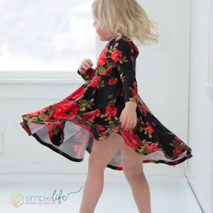 May include: A young girl wearing a black and red floral dress with long sleeves and a twirling skirt. She is wearing gold sandals and is standing on a white floor.