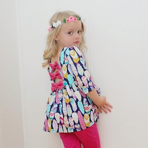 May include: A young girl wearing a colourful feather print dress with pink bows on the back. She is wearing a flower crown and pink trousers.