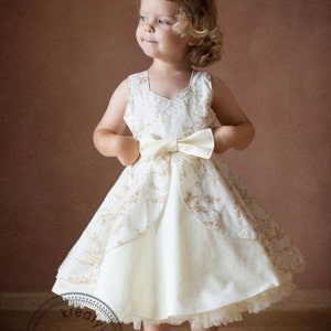 May include: A young girl wearing a white dress with gold embroidery and a large white bow. The dress has a layered skirt and is worn with white shoes.