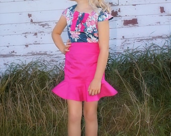 Puede incluir: Una niña pequeña con una falda rosa y una blusa con estampado floral. La falda tiene un dobladillo con volantes y la blusa tiene un lazo en el escote. Está de pie en una zona cubierta de hierba.