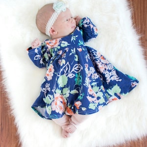 May include: A baby wearing a blue floral dress with long sleeves and a white headband with a light blue flower. The baby is lying on a white fluffy rug.